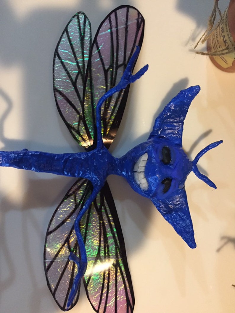 May include: A blue dragonfly sculpture with iridescent wings. The dragonfly has a toothy grin and large, pointed ears.