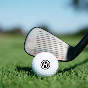 K&ouml;nnte beinhalten: Ein wei&szlig;er Golfball mit einem schwarzen "H" in einem Kreis darauf, der auf gr&uuml;nem Gras liegt. Ein schwarzer Golfschl&auml;ger liegt auf dem Ball.