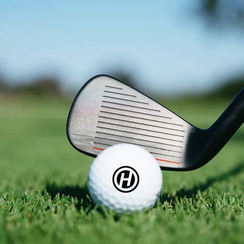 K&ouml;nnte beinhalten: Ein wei&szlig;er Golfball mit einem schwarzen "H" in einem Kreis darauf, der auf gr&uuml;nem Gras liegt. Ein schwarzer Golfschl&auml;ger liegt auf dem Ball.