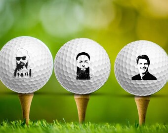 Könnte beinhalten: Drei weiße Golfbälle mit schwarz-weißen Bildern von Männergesichtern darauf gedruckt. Die Golfbälle liegen auf Golftees.