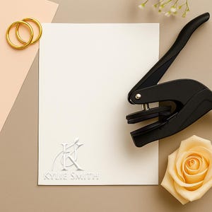 May include: A cream-colored card with embossed initials and the name "KYLIE SMITH" is next to a black paper punch, gold rings, and a peach rose. The items are arranged on a neutral-toned surface.