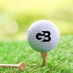 K&ouml;nnte beinhalten: Ein wei&szlig;er Golfball mit den Buchstaben "GB" in Schwarz darauf gedruckt. Der Ball liegt auf einem Abschlag in einem gr&uuml;nen Grasbereich.