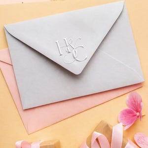 May include: A light gray envelope featuring a silver embossed monogram with the letters H and C intertwined. The envelope is positioned over a light pink envelope, with a pink flower petal and light pink ribbon visible in the lower corner.