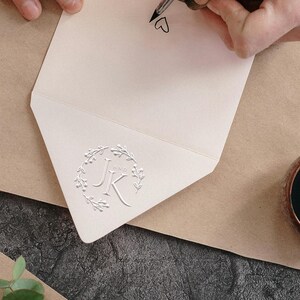 Op de afbeelding: Een open, crèmekleurige envelop met een in reliëf aangebracht bloemenkransontwerp rond de letters "JK" en "AND". Een hand schrijft een hartsymbool met een pen op de envelop. De envelop ligt op een bruin oppervlak.