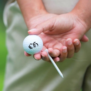 K&ouml;nnte beinhalten: Ein wei&szlig;er Golfball mit der Nummer "C5" darauf, in einer Hand mit einem Abschlagtee gehalten.