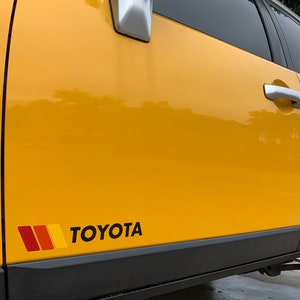 Op de afbeelding: Een gele Toyota FJ Cruiser met een zwarte en rode streep sticker en het woord "TOYOTA" in zwarte letters aan de zijkant van het voertuig.