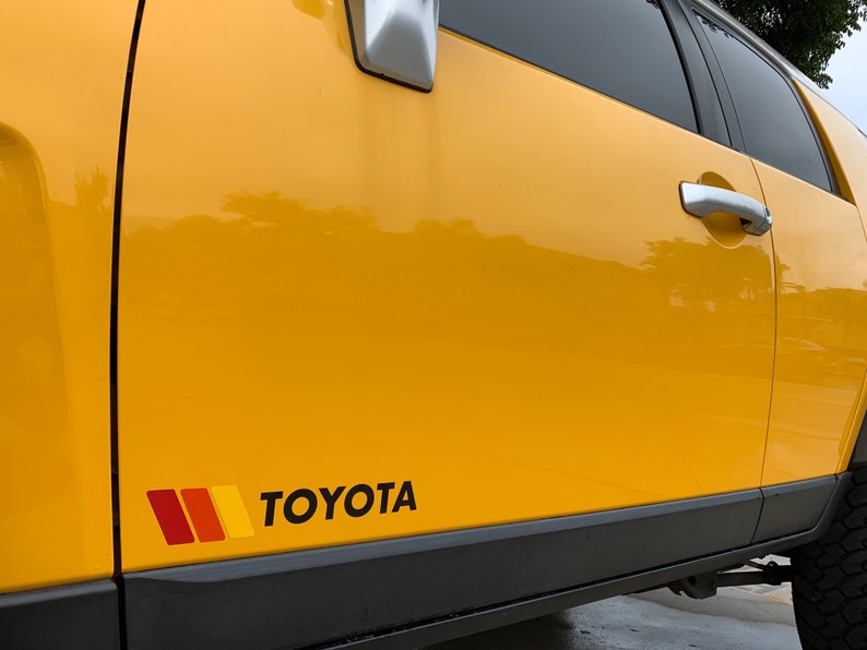 Op de afbeelding: Een gele Toyota FJ Cruiser met een zwarte en rode streep sticker en het woord "TOYOTA" in zwarte letters aan de zijkant van het voertuig.