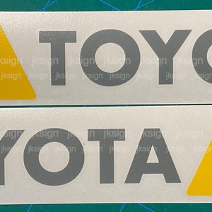 Op de afbeelding: Twee witte stickers met het woord "TOYOTA" in grijze letters. De stickers hebben drie strepen in rood, oranje en geel.