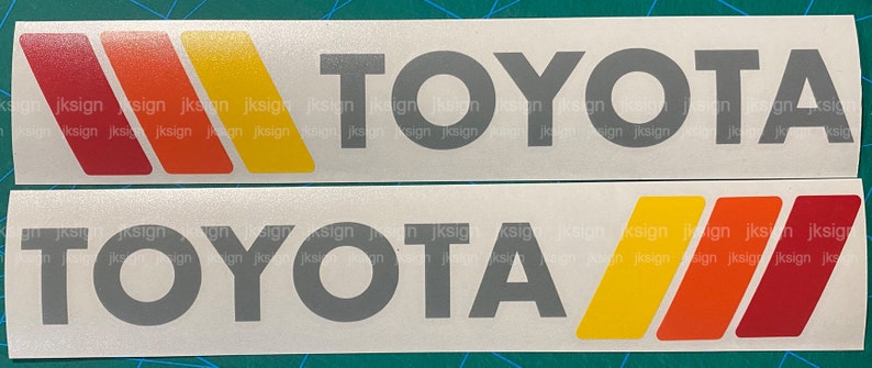 Op de afbeelding: Twee witte stickers met het woord "TOYOTA" in grijze letters. De stickers hebben drie strepen in rood, oranje en geel.