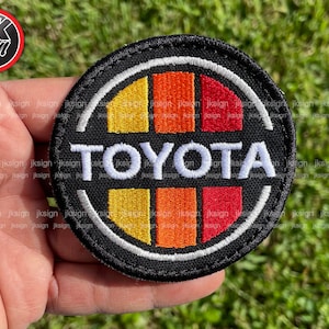 May include: A black embroidered patch with a white circle around a red, orange, and yellow design. The word "TOYOTA" is embroidered in white letters in the center of the patch.