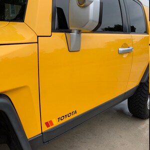 Op de afbeelding: Gele Toyota SUV met zwarte bekleding en een zijspiegel. Het voertuig heeft een zwarte en rode streepsticker met het woord "TOYOTA" aan de zijkant.