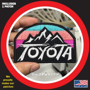 May include: A black embroidered patch with a mountain range and the word "TOYOTA" in white. The mountains are set against a sunset background with pink, orange, and blue stripes. The patch is 3.8 inches wide and 2.5 inches high.