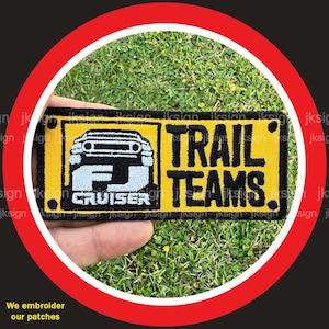 May include: Yellow and black embroidered patch with a white outline. The patch features a cartoon image of a Toyota FJ Cruiser with the text "FJ Cruiser" and "TRAIL TEAMS."