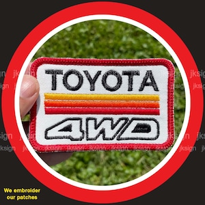 May include: White embroidered patch with red stitching. The patch features the text "TOYOTA" in black lettering with three red, orange, and yellow stripes above it. Below the text is "4WD" in black lettering.