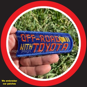 May include: A blue embroidered patch with the text "OFF-ROAD WITH TOYOTA" in orange and yellow thread. The patch has a yellow arrow pointing to the right.