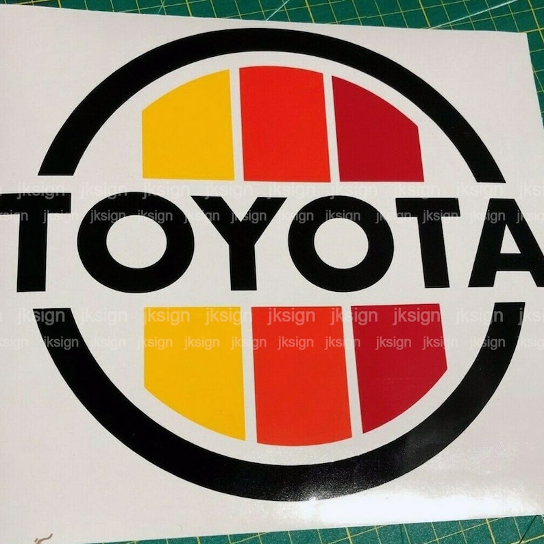 Peut inclure: Un logo circulaire noir et blanc avec le mot "TOYOTA" en lettres noires en gras. Le cercle a un design rouge, orange et jaune &agrave; l'int&eacute;rieur.