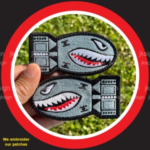 May include: Two embroidered patches in the shape of a cartoon bomb with a shark-like design. The patches are gray with black borders, white teeth, and red accents. The text "We embroider our patches" is at the bottom.
