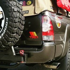 Peut inclure: Un pick-up Toyota Tacoma noir avec une capote en toile beige. Le camion a une roue de secours mont&eacute;e &agrave; l'arri&egrave;re, un attelage de remorquage rouge et un autocollant "TRO" rouge, jaune et orange &agrave; l'arri&egrave;re.