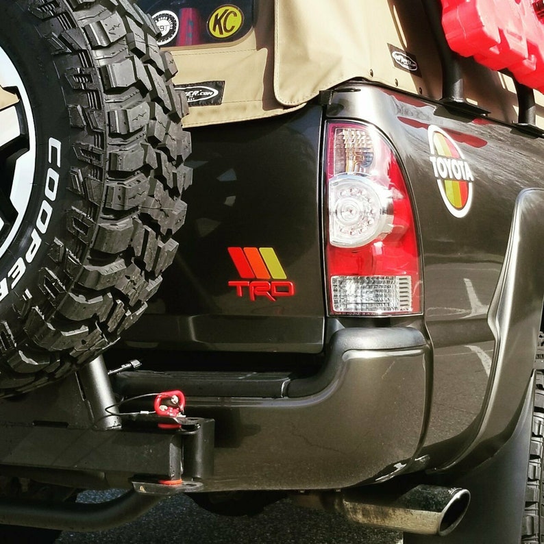 Peut inclure: Un pick-up Toyota Tacoma noir avec une capote en toile beige. Le camion a une roue de secours mont&eacute;e &agrave; l'arri&egrave;re, un attelage de remorquage rouge et un autocollant "TRO" rouge, jaune et orange &agrave; l'arri&egrave;re.