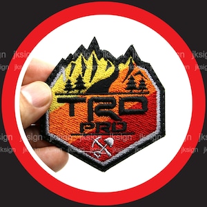 May include: A black, red, and yellow embroidered patch with the words "TRD PRO" and a mountain range with trees. A pickaxe and shovel are below the text.
