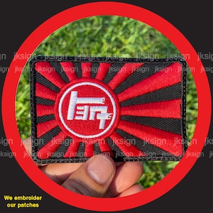 May include: A red and black embroidered patch with a white Japanese symbol in the center. The symbol is surrounded by a red circle and the background is a red and black rising sun design.