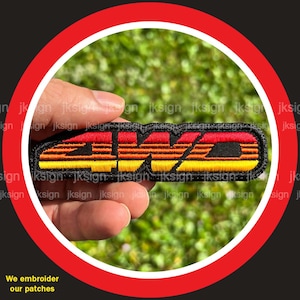 May include: A black embroidered patch with the word "4WD" in white letters with red, orange, and yellow stripes. The patch is held in a hand against a blurred green background.