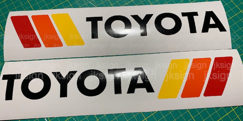 Op de afbeelding: Twee witte stickers met het woord "TOYOTA" in zwarte letters. De bovenste sticker heeft drie strepen in rood, oranje en geel. De onderste sticker heeft drie strepen in geel, oranje en rood.