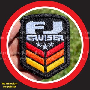 May include: Embroidered patch with a black background, white lettering that says "FJ CRUISER" with three white stars, and three stripes in red, orange, and yellow.