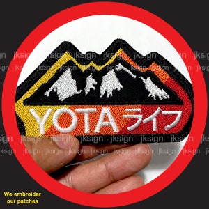 May include: A black and white embroidered patch with a mountain range design. The mountains are outlined in yellow and orange. The text "YOTA ライフ" is embroidered in white on a red background.
