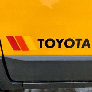 Op de afbeelding: Een gele auto met een zwart "TOYOTA" logo en een rode, oranje en gele streep sticker aan de zijkant.