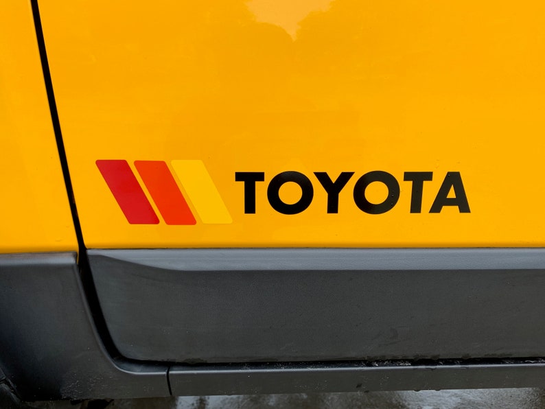 Op de afbeelding: Een gele auto met een zwart "TOYOTA" logo en een rode, oranje en gele streep sticker aan de zijkant.
