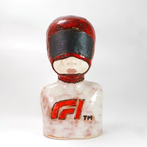 Puede incluir: Un busto de cerámica de una persona que lleva un casco rojo con una visera negra y una camisa blanca con un logotipo "F1" rojo y el texto "TM".