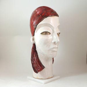 May include: A ceramic sculpture of a stylized human head. The head is primarily white with red hair and accents. The eyes are detailed with gold-colored embellishments. The sculpture is mounted on a white square base.