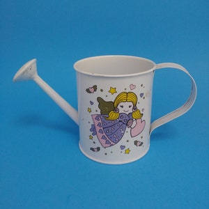 Puede incluir: Regadera de metal blanco con boquilla larga y asa. La regadera presenta un diseño de ángel de dibujos animados con cabello rubio, alas y un corazón. El fondo es azul claro.