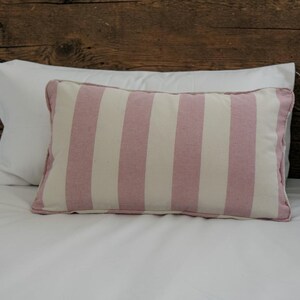 May include: A rectangular throw cushion featuring vertical stripes in muted dusty pink and off-white. The cushion has a piped edge trim in the same dusty pink colour, creating a defined border around the rectangular shape of the cushion.