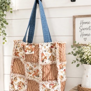 May include: A quilted tote bag featuring a patchwork design of autumn-themed fabric squares with foxes, mushrooms, and acorns, alternating with brown and tan striped fabric. The bag has frayed seams and two blue denim shoulder straps.