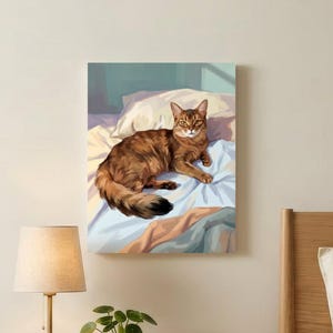 May include: A rectangular wall art print featuring a brown tabby cat resting on white and blue bedding. The artwork is displayed above a light wood nightstand that holds a lamp with a beige shade, a small green potted plant, and a ceramic mug.