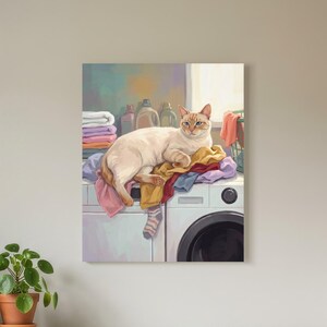 May include: A rectangular wall art print depicting a cream-colored cat resting on a pile of colorful folded laundry atop a white washing machine. The artwork features soft pastel tones, including shades of pink, blue, and yellow, with a single striped sock hanging over the edge of the machine.