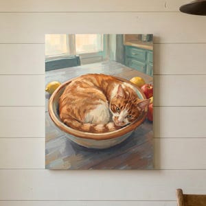 May include: A framed wall art print depicting an orange tabby cat curled up inside a ceramic bowl on a wooden table. The scene includes yellow lemons and red apples placed around the bowl, rendered in a painterly style with warm lighting.