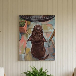 May include: A rectangular wall art print featuring a brown dog sitting on a colorful stone patio, looking upward with a small wisp of steam rising above its head. The artwork is displayed on a vertical white paneled wall between two rattan chairs.