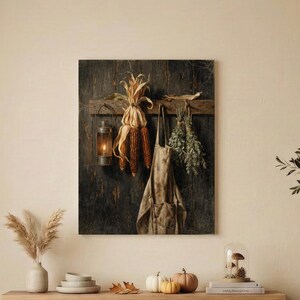 May include: A rectangular wall art print featuring a dark wooden background with a hanging lantern, dried corn husks, a beige apron, and a bundle of green herbs. The artwork is displayed above a long wooden console table in a neutral room.
