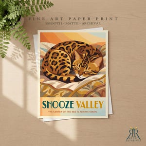 May include: A fine art paper print featuring a curled-up leopard-spotted cat on a patterned blanket. The text reads SNOOZE VALLEY and THE CENTER OF THE BED IS ALWAYS TAKEN. The illustration uses warm orange, yellow, and brown tones.