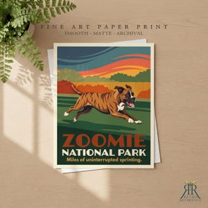 May include: A fine art paper print featuring a brindle boxer dog running across a green field. The text reads ZOOMIE NATIONAL PARK and Miles of uninterrupted sprinting. The background shows a sunset sky with orange and blue clouds.