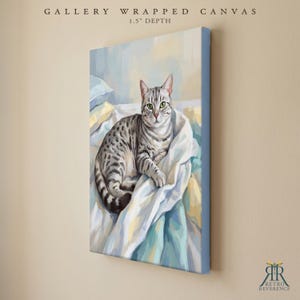 May include: A gallery wrapped canvas with a 1.5 inch depth featuring a painting of a grey tabby cat with green eyes resting on white and blue bedding. The text GALLERY WRAPPED CANVAS 1.5" DEPTH appears at the top, with a logo at the bottom.
