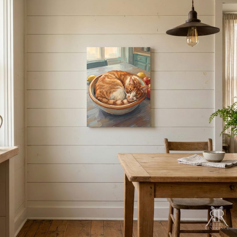 May include: A framed wall art print depicting an orange tabby cat curled up inside a ceramic bowl on a wooden table. The scene includes yellow lemons and red apples placed around the bowl, rendered in a painterly style with warm lighting.