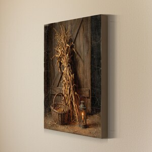 May include: A rectangular canvas print featuring a bundle of dried corn stalks leaning against a weathered brown wooden door. A woven wicker basket sits to the left, and a metal lantern with a glowing candle is positioned on the ground.