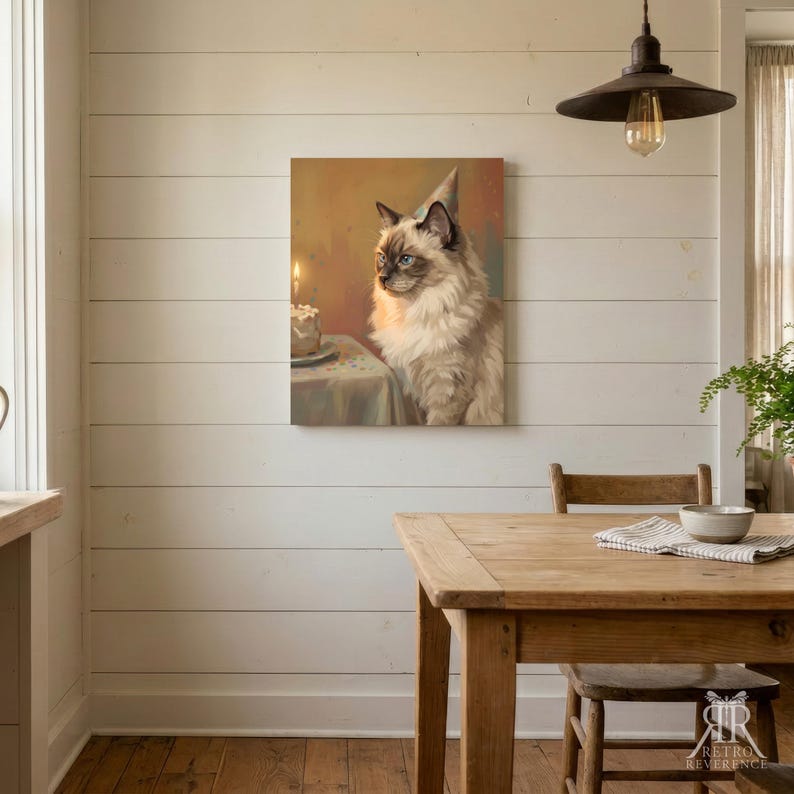 May include: A square wall art print featuring a fluffy, long-haired cat with blue eyes and dark points, wearing a small party hat. The cat sits beside a table with a lit birthday cake. The background is a warm, soft-toned, painterly scene.
