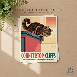 May include: A fine art paper print featuring a brown tabby cat perched on the edge of a red kitchen counter. The text reads: FINE ART PAPER PRINT, COUNTERTOP CLIFFS, Not recommended. Wildy popular anyway. The print has a smooth matte finish.