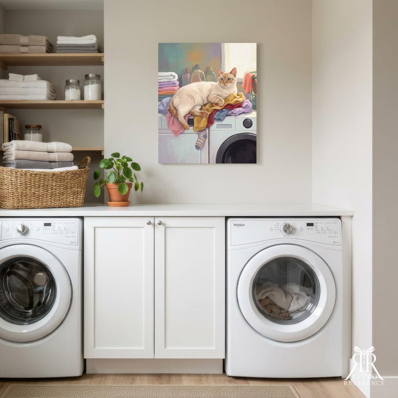 May include: A rectangular wall art print depicting a cream-colored cat resting on a pile of colorful folded laundry atop a white washing machine. The artwork features soft pastel tones, including shades of pink, blue, and yellow, with a single striped sock hanging over the edge of the machine.