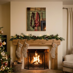 May include: A rustic fireplace mantel made of light brown wood features three beige knit stockings hanging from the edge. A green garland with red berries and warm lights sits atop the mantel. A painting of a wall with hanging items is above.